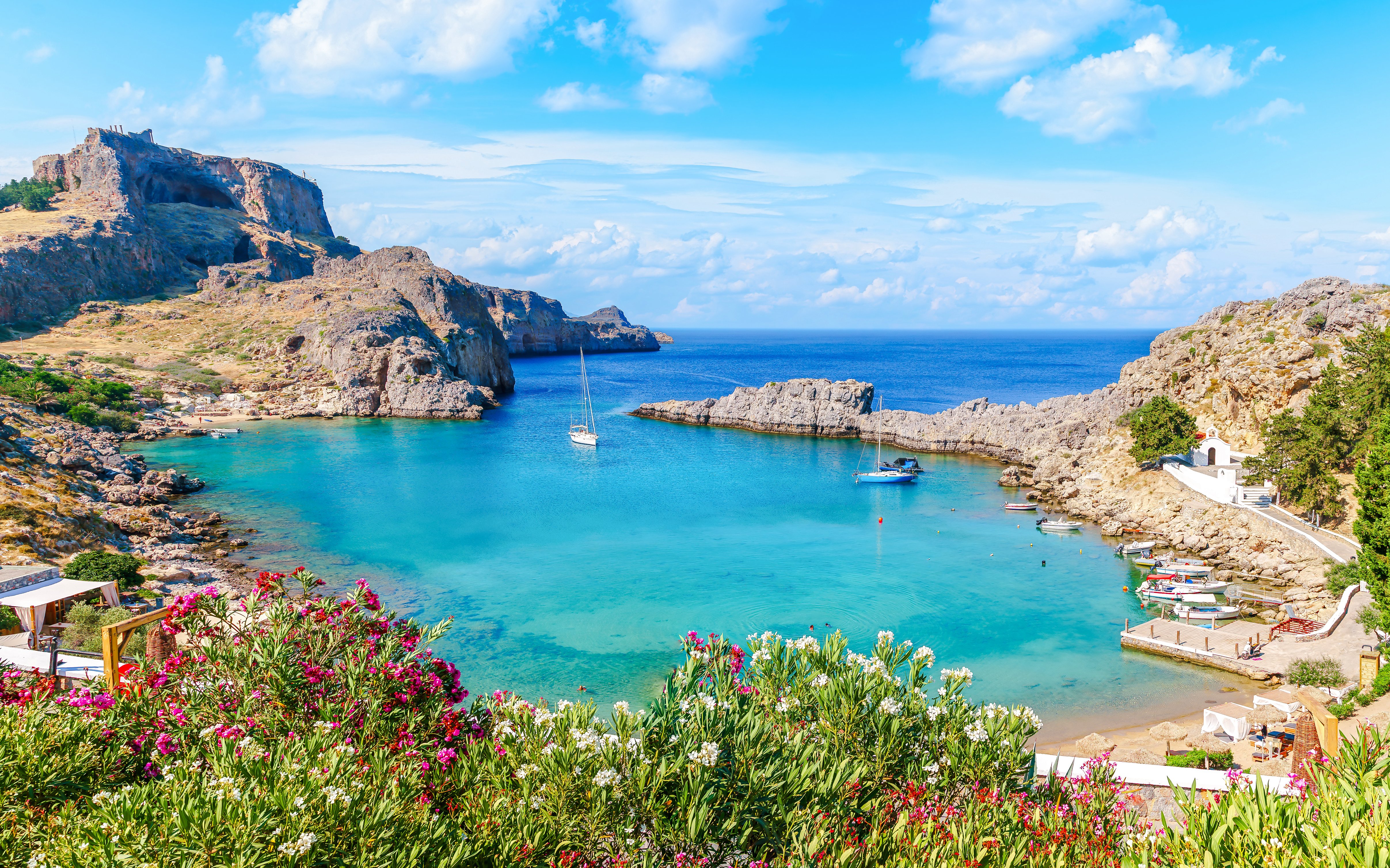 Sailboats in the turquoise waters of Saint Paul Bay, Rhodes, Greece, with rocky cliffs and vibrant flowers.