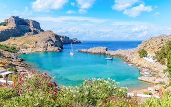 Sailboats in the turquoise waters of Saint Paul Bay, Rhodes, Greece, with rocky cliffs and vibrant flowers.