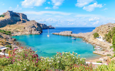 Sailboats in the turquoise waters of Saint Paul Bay, Rhodes, Greece, with rocky cliffs and vibrant flowers.