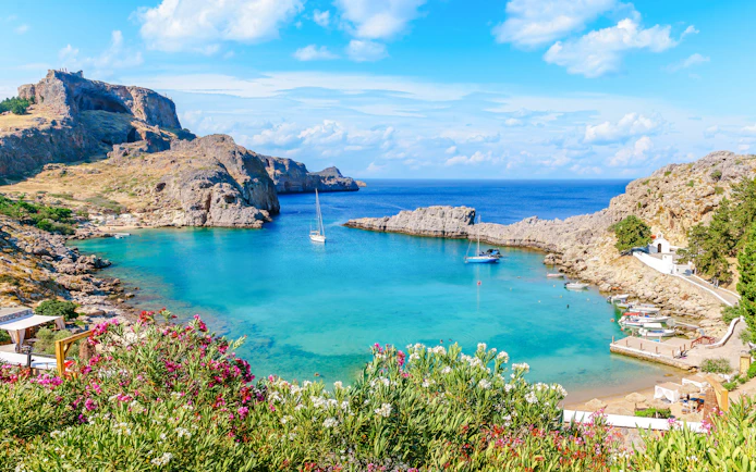 Sailboats in the turquoise waters of Saint Paul Bay, Rhodes, Greece, with rocky cliffs and vibrant flowers.