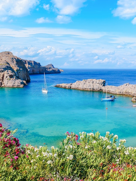 Sailboats in the turquoise waters of Saint Paul Bay, Rhodes, Greece, with rocky cliffs and vibrant flowers.