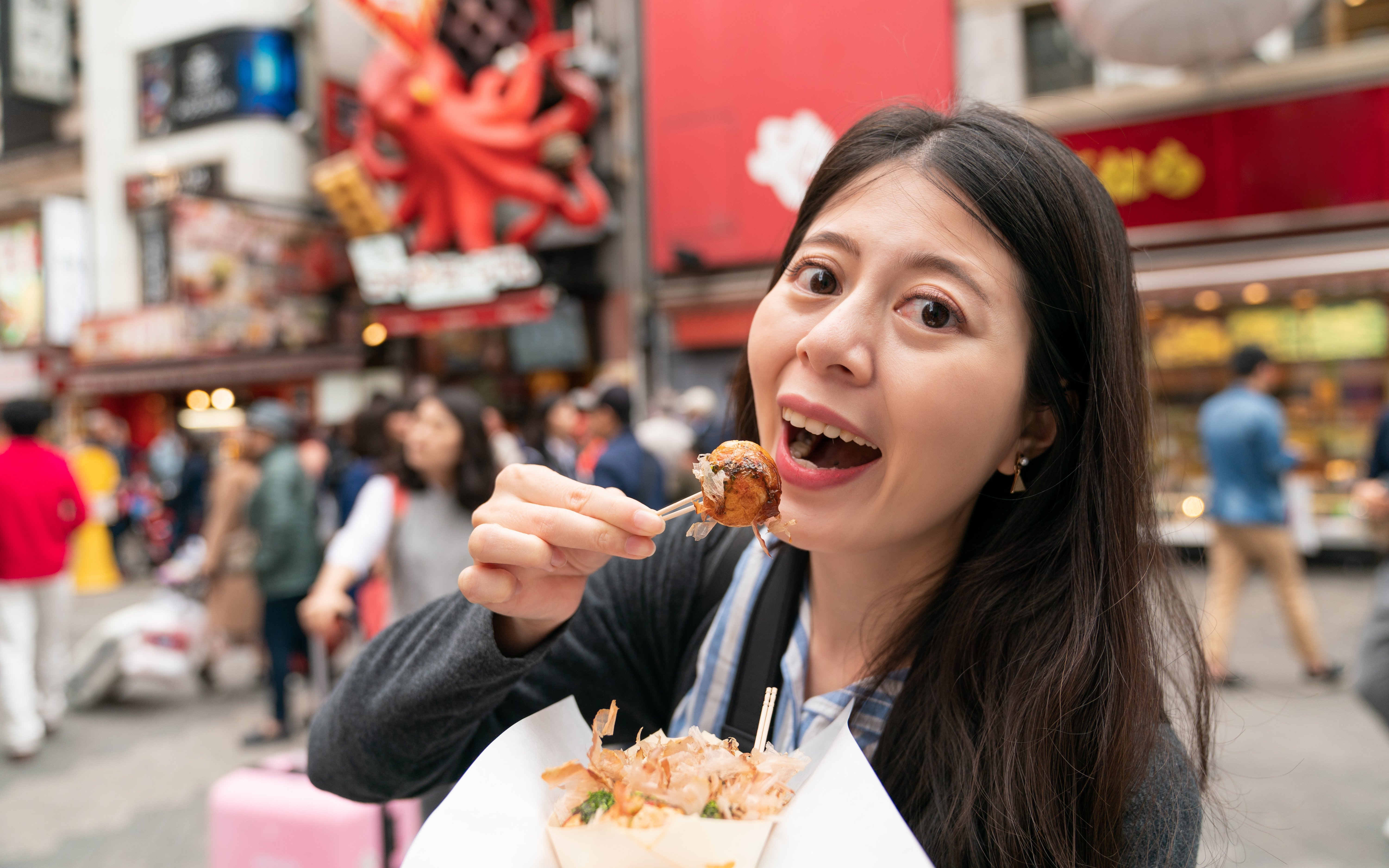 Asian woman enjoying takoyaki on a bustling street in Japan.