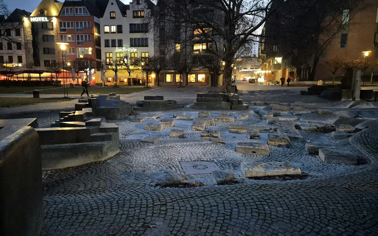 Cobblestone square with stone sculptures and lit buildings in Cologne at dusk.