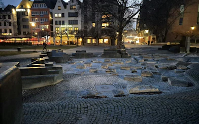 Cobblestone square with stone sculptures and lit buildings in Cologne at dusk.