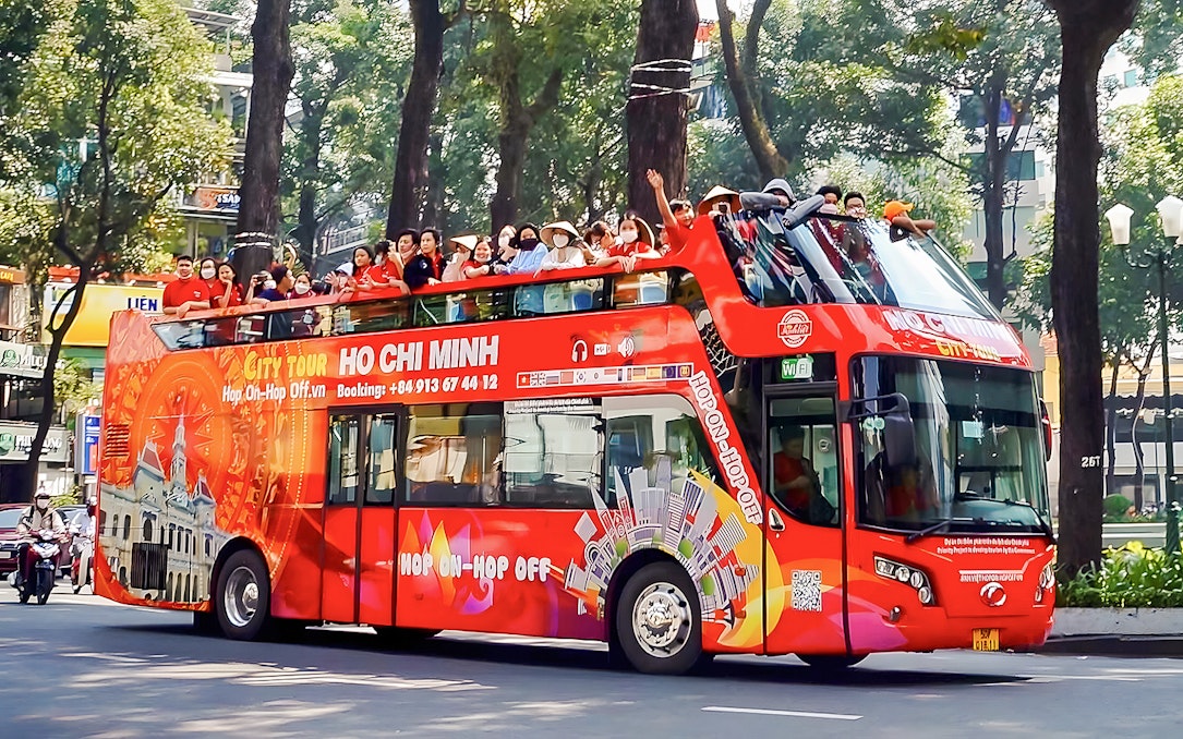 Hop-on Hop-off bus with tourists in Ho Chi Minh City, Vietnam, passing tree-lined street.