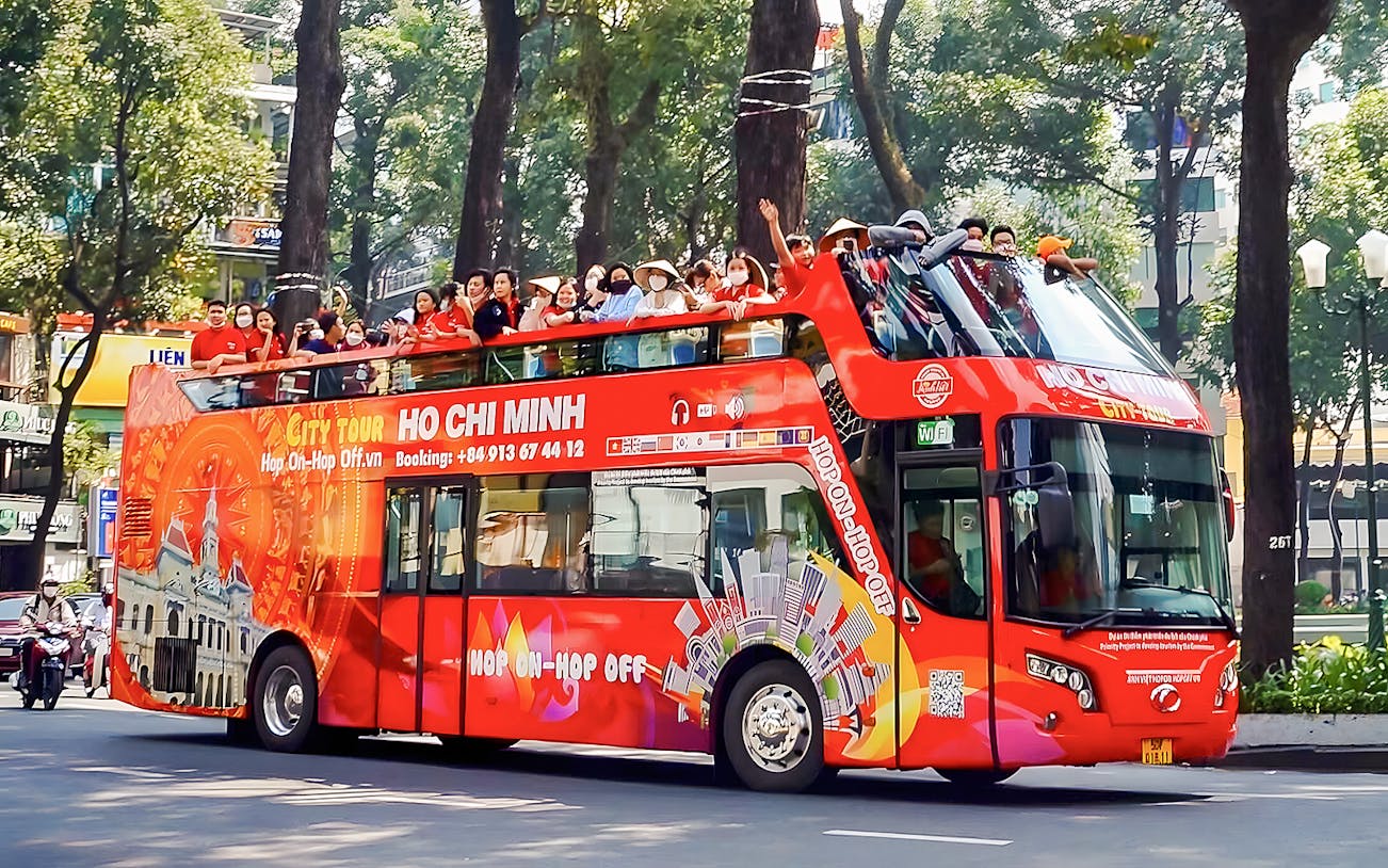 Hop-on Hop-off bus with tourists in Ho Chi Minh City, Vietnam, passing tree-lined street.