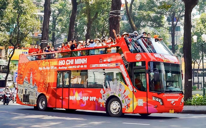 Hop-on Hop-off bus with tourists in Ho Chi Minh City, Vietnam, passing tree-lined street.