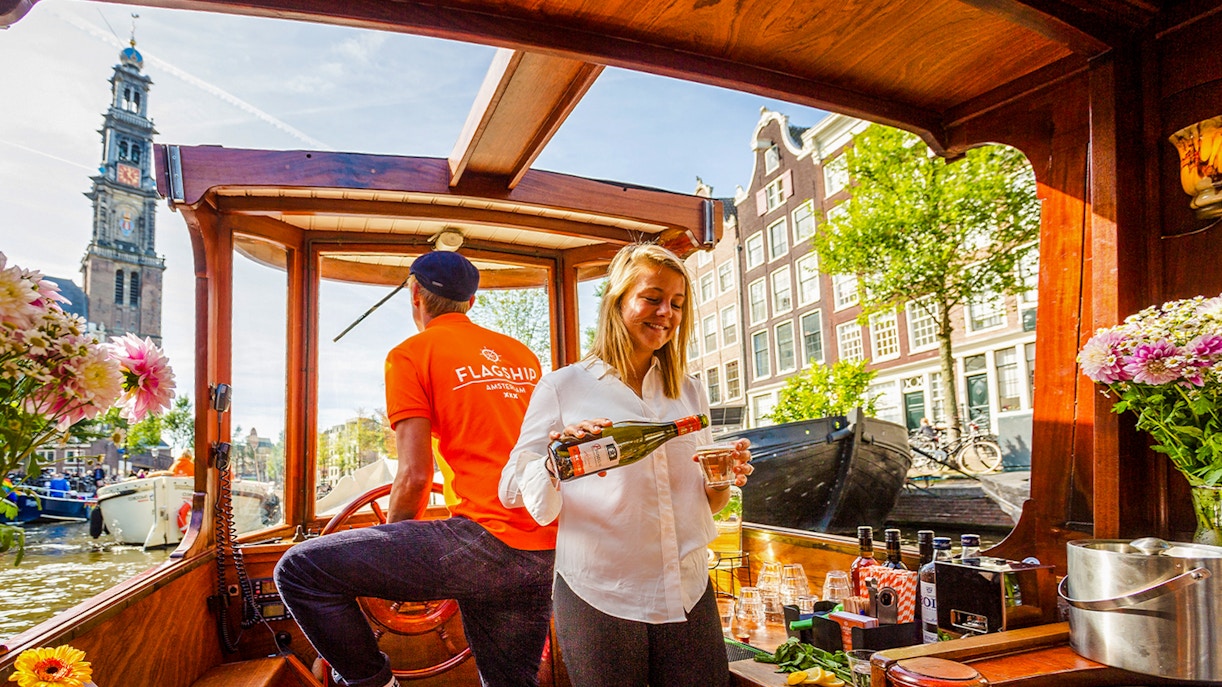 Guide serving wine on Amsterdam canal cruise boat with Westerkerk in background.