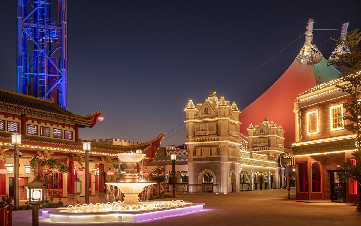 Six Flags Qiddiya City at night with illuminated buildings and a central fountain.