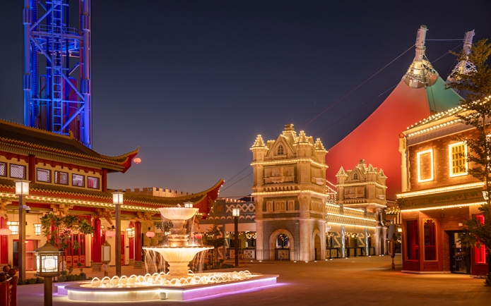 Six Flags Qiddiya City at night with illuminated buildings and a central fountain.