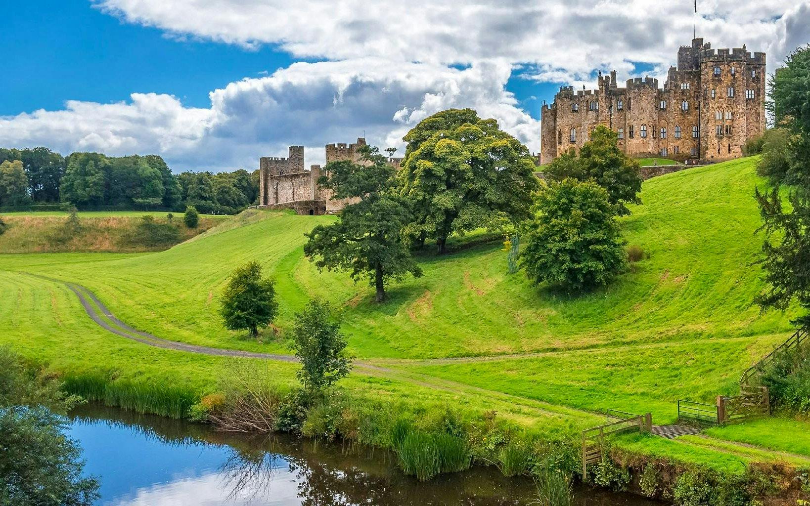 Alnwick Castle exteriors with lush green landscape and reflective pond.