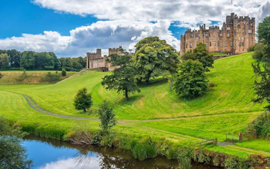 Alnwick Castle exteriors with lush green landscape and reflective pond.