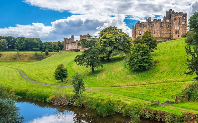 Alnwick Castle exteriors with lush green landscape and reflective pond.