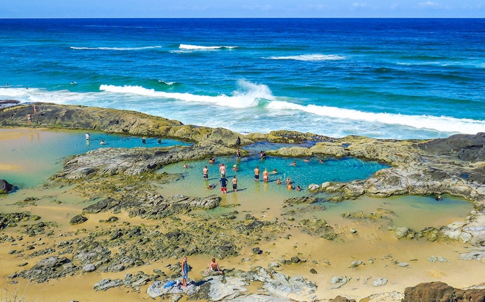 People swimming in natural rock pools on Fraser Island, K'gari, with ocean waves in the background.