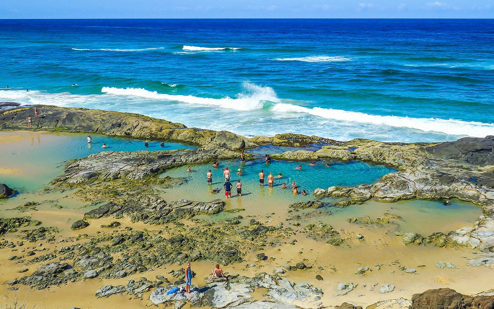 People swimming in natural rock pools on Fraser Island, K'gari, with ocean waves in the background.