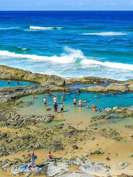 People swimming in natural rock pools on Fraser Island, K'gari, with ocean waves in the background.