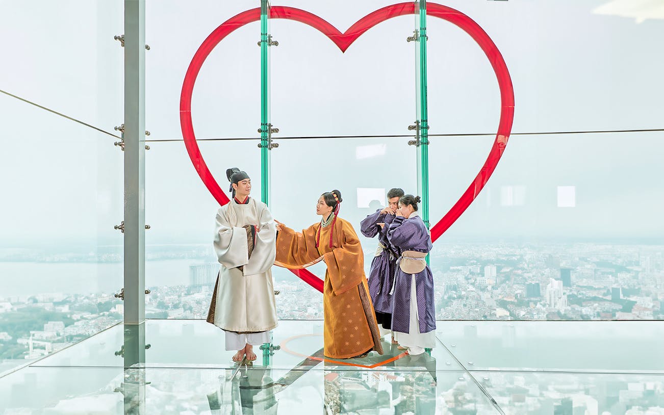 Visitors in traditional attire on glass floor at Hanoi Sky Lotte Observation Deck with city view.
