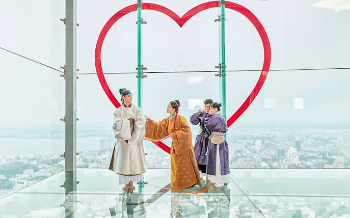 Visitors in traditional attire on glass floor at Hanoi Sky Lotte Observation Deck with city view.