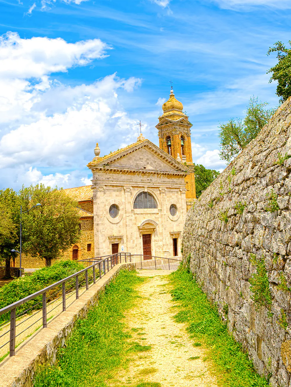 Historic church with bell tower in Montalcino, Italy, surrounded by stone walls and greenery.
