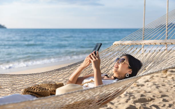 Lady relaxing in a hammock on a beach using a cellphone.