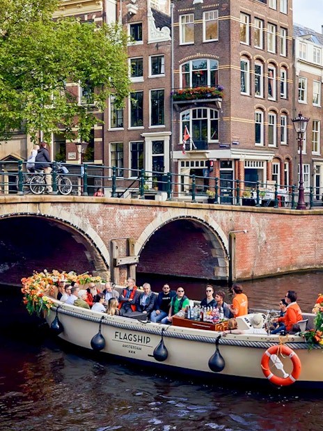 Boat with flowers and passengers on Amsterdam canal near brick bridge.