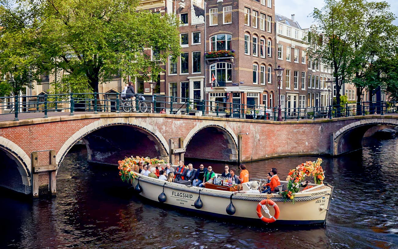 Boat with flowers and passengers on Amsterdam canal near brick bridge.