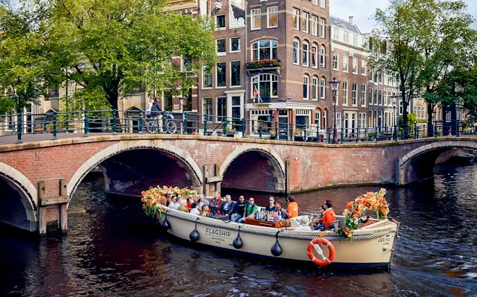 Boat with flowers and passengers on Amsterdam canal near brick bridge.