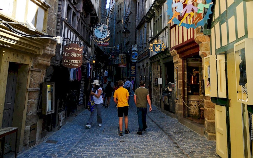 Narrow cobblestone street in Mont-Saint-Michel with shops and tourists walking.