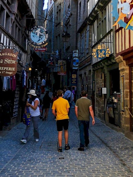 Narrow cobblestone street in Mont-Saint-Michel with shops and tourists walking.