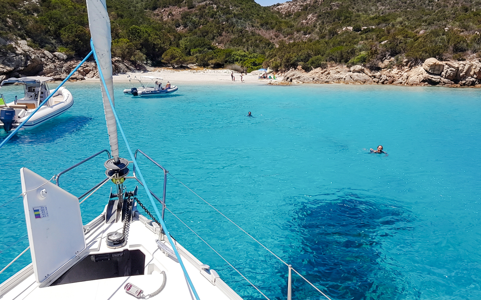Sailboat approaching a beach in La Maddalena Archipelago with swimmers in clear blue water.