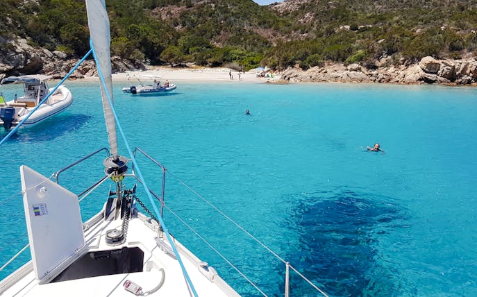 Sailboat approaching a beach in La Maddalena Archipelago with swimmers in clear blue water.