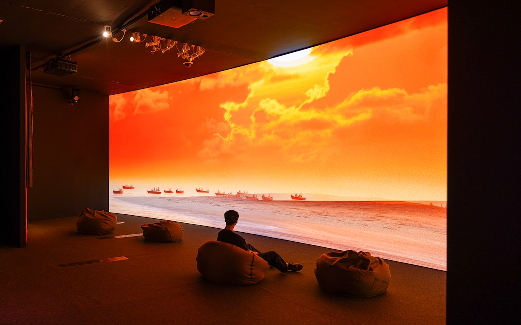 Man in dark room watches large screen of boats on sunset sea.