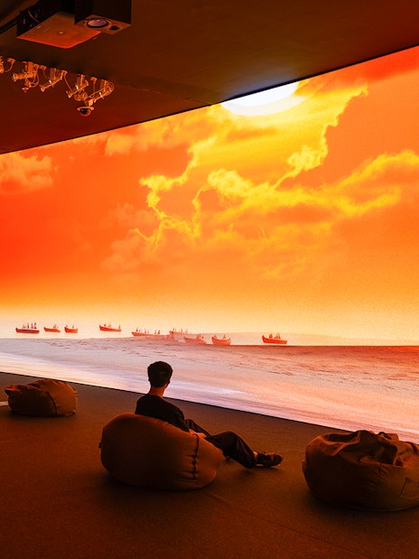 Man in dark room watches large screen of boats on sunset sea.