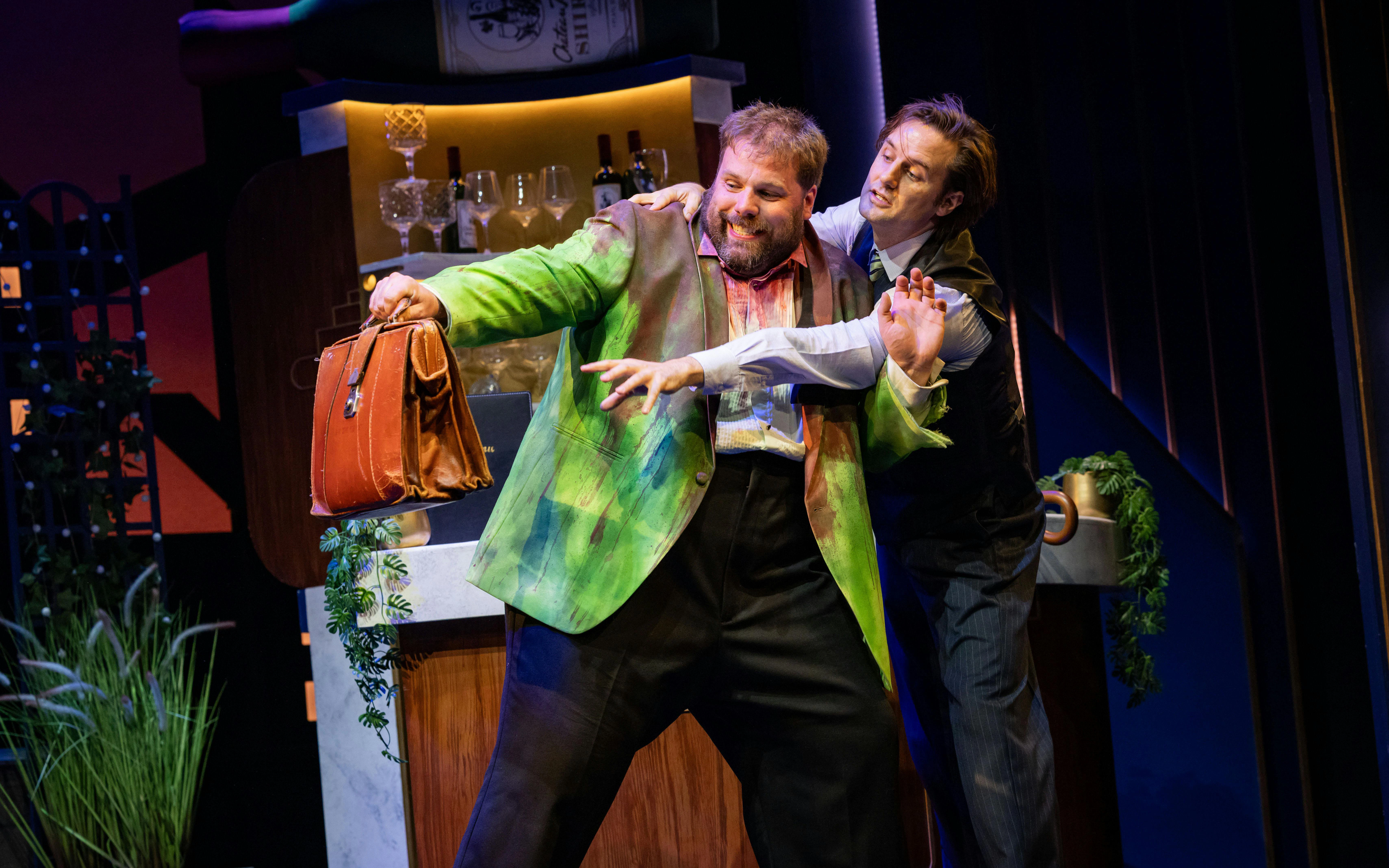 Actors in a comedic scene from "The Comedy About Spies" on stage, one holding a briefcase.