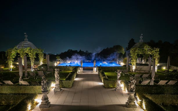 Exteriors of QC Terme Garda at night with illuminated pool and garden statues.