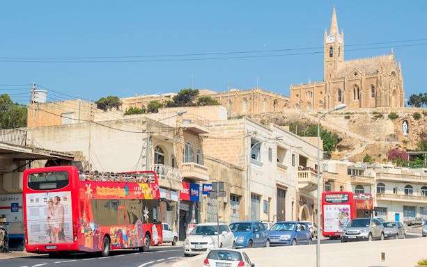 Red double-decker bus on Gozo Hop On Hop Off tour passing Ta' Pinu Basilica, Malta.