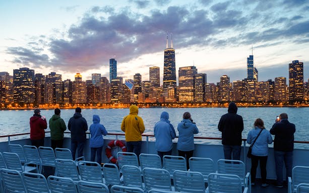 People on a boat deck viewing Chicago skyline at dusk, water in foreground, cloudy sky above.