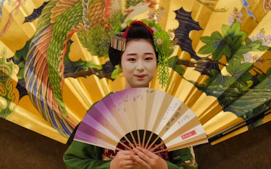 Geisha holding a fan in front of a decorative Japanese screen.