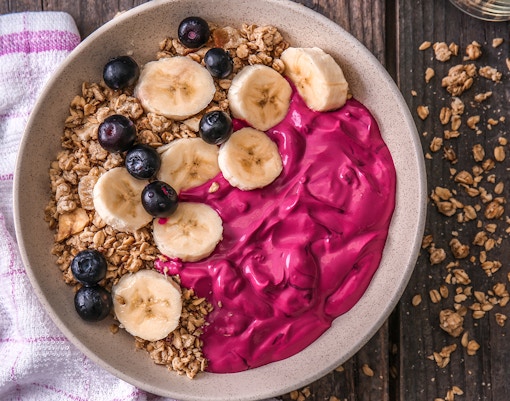 Granola with banana slices, blueberries, and pink yogurt in a bowl on a wooden table.