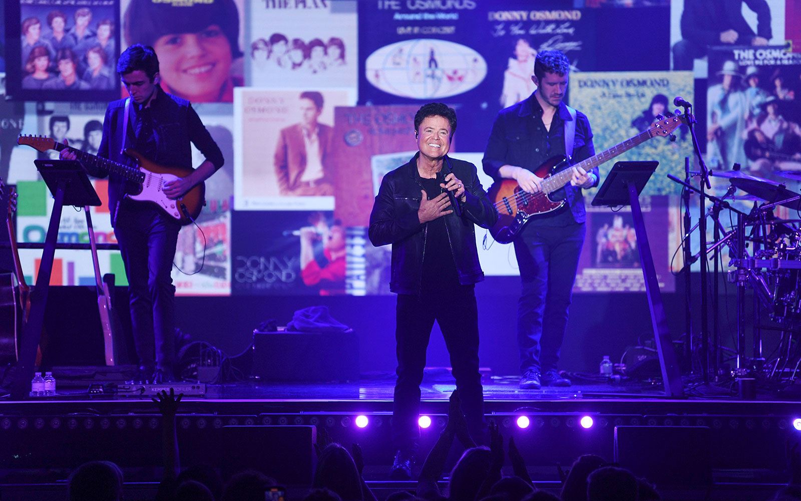 Singer performing on stage with musicians and album covers in the background.