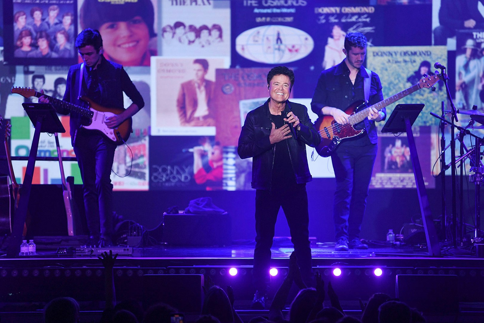 Singer performing on stage with musicians and album covers in the background.