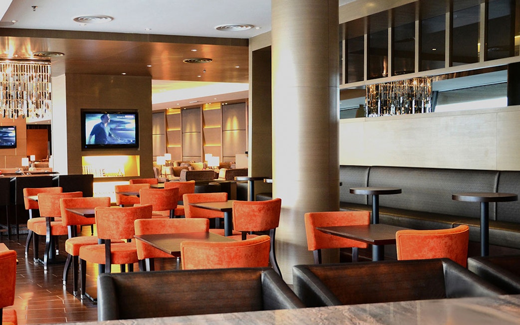 Plaza Premium First lounge seating area with orange chairs and a TV, Satellite Terminal.