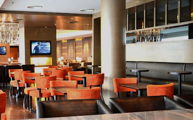 Plaza Premium First lounge seating area with orange chairs and a TV, Satellite Terminal.
