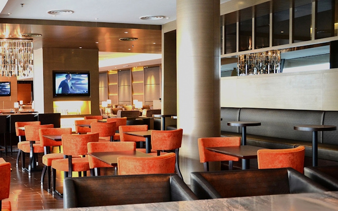 Plaza Premium First lounge seating area with orange chairs and a TV, Satellite Terminal.