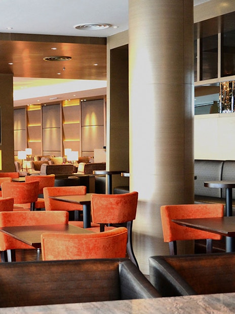 Plaza Premium First lounge seating area with orange chairs and a TV, Satellite Terminal.