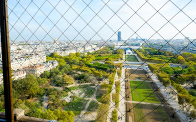 View of Paris from Eiffel Tower summit through safety grid, showcasing Champ de Mars and cityscape.