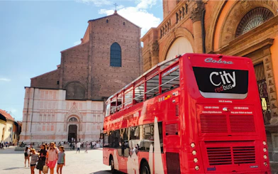 Red double-decker bus near historic church in Bologna, Italy, with tourists nearby.