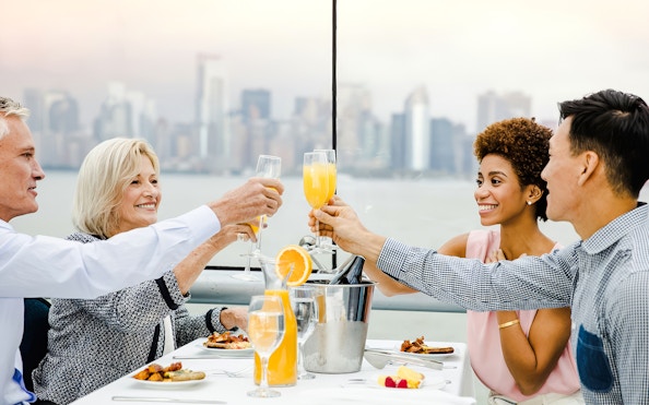 Guests toasting with mimosas on a luxury glass boat brunch cruise by Bateaux, city skyline in background.