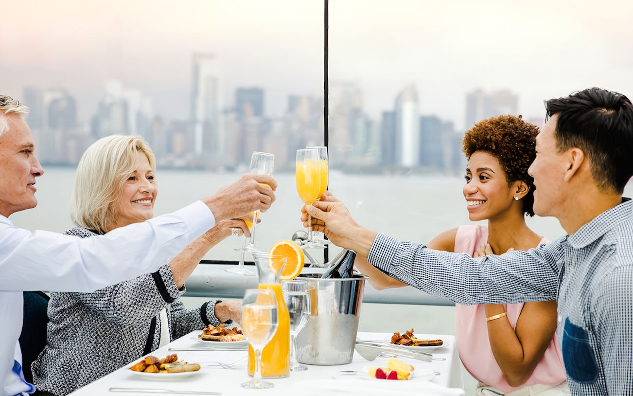 Guests toasting with mimosas on a luxury glass boat brunch cruise by Bateaux, city skyline in background.
