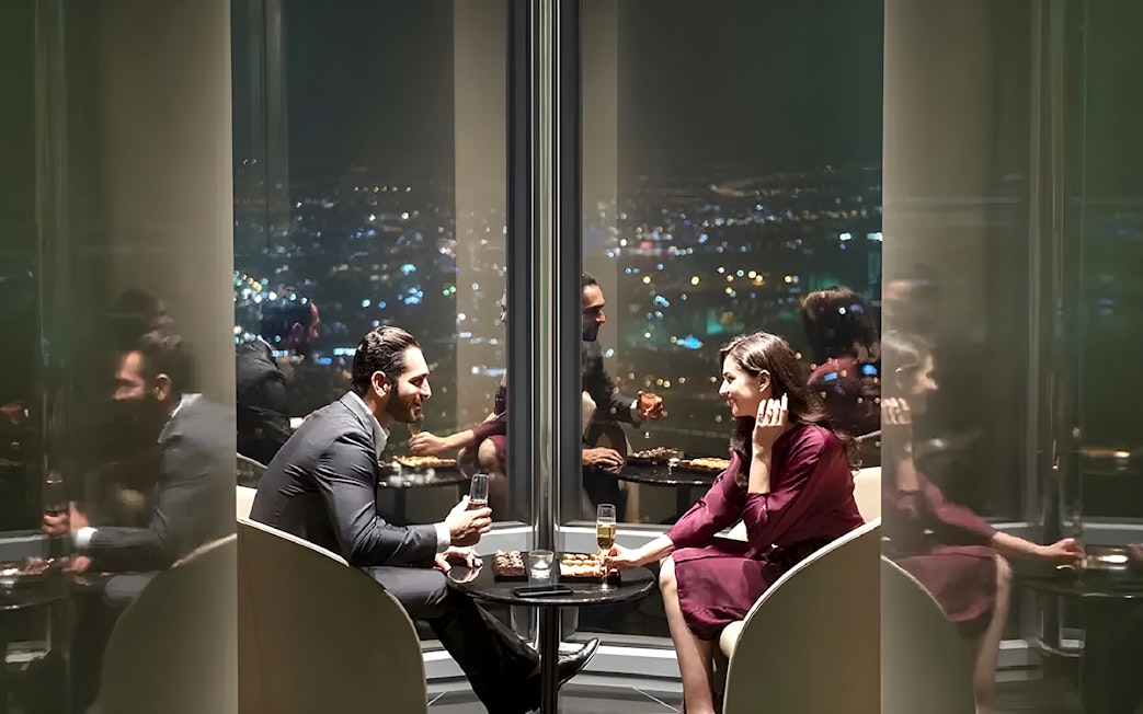 Couple enjoying drinks at Burj Khalifa's At the Top Sky Lounge, overlooking city lights.
